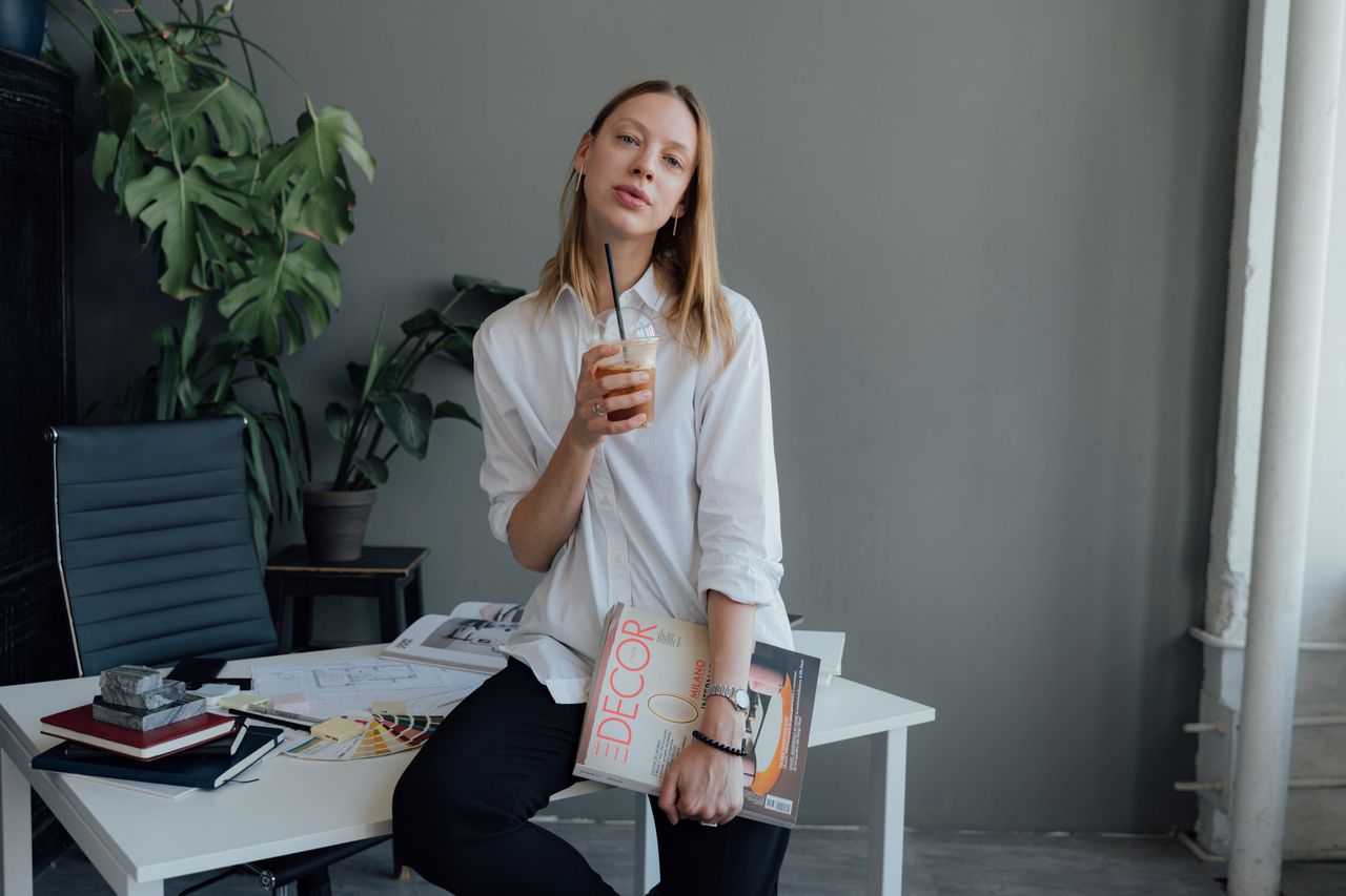 Elegant woman in a modern office sipping a drink, showcasing fashion and style.