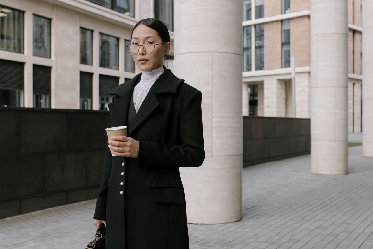 our-story Asian businesswoman holding a coffee cup and standing outdoors in a modern urban area.