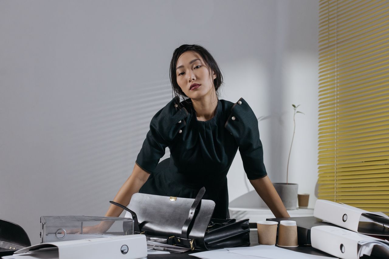 heros-img Asian woman in black dress posing confidently in a stylish office setting with modern decor.